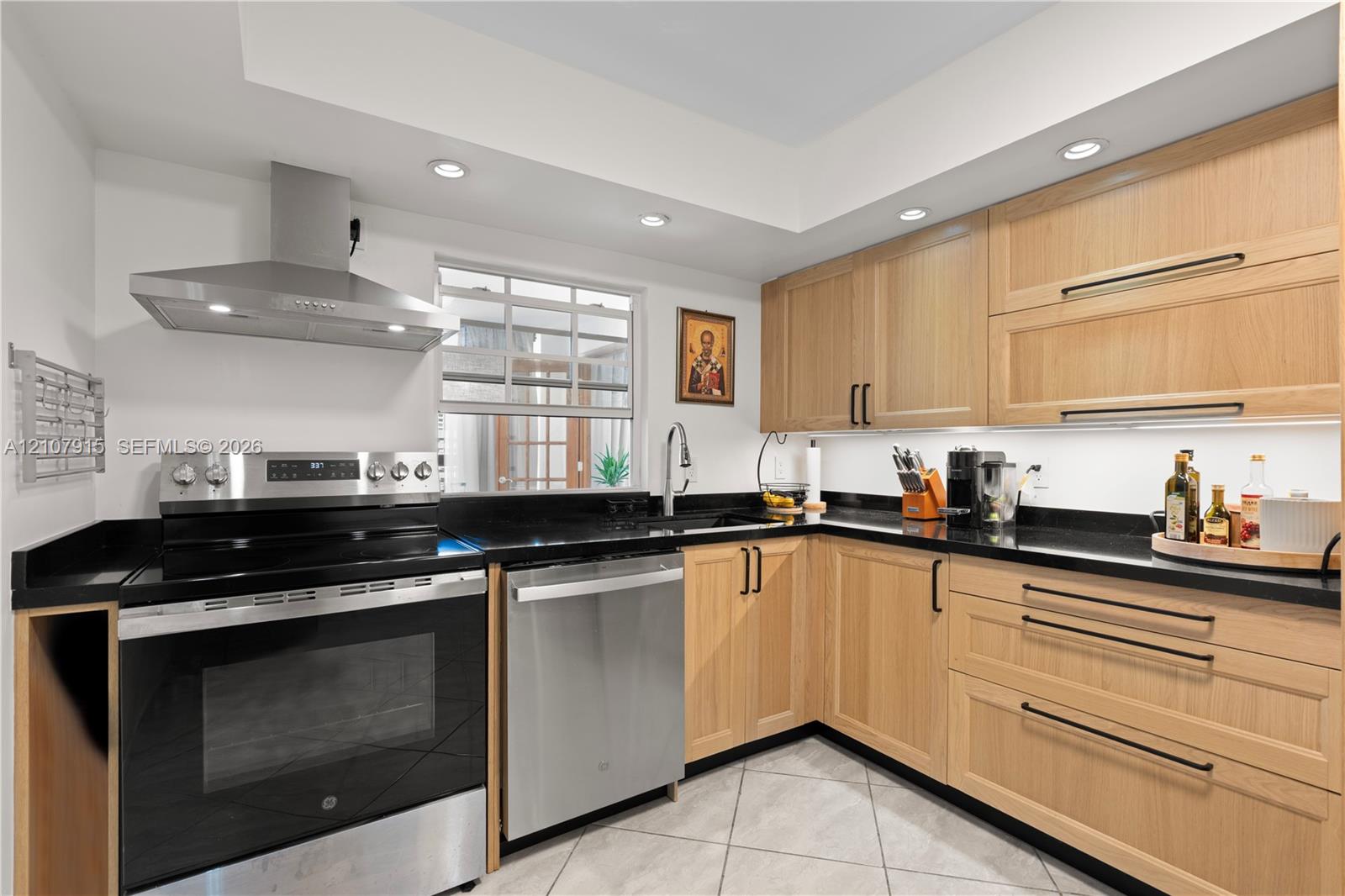 Kitchen featuring light wood-finish cabinetry, dark solid-surface countertops, stainless steel appliances, a recessed range hood, and tile flooring