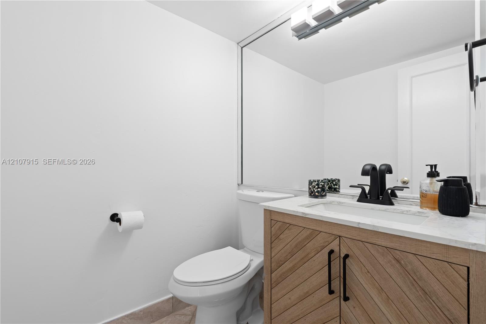 Bathroom vanity with wood-finish cabinetry featuring a diagonal grain pattern, stone-top counter with an integrated basin, matte black faucet hardware, and a full-width wall mirror
