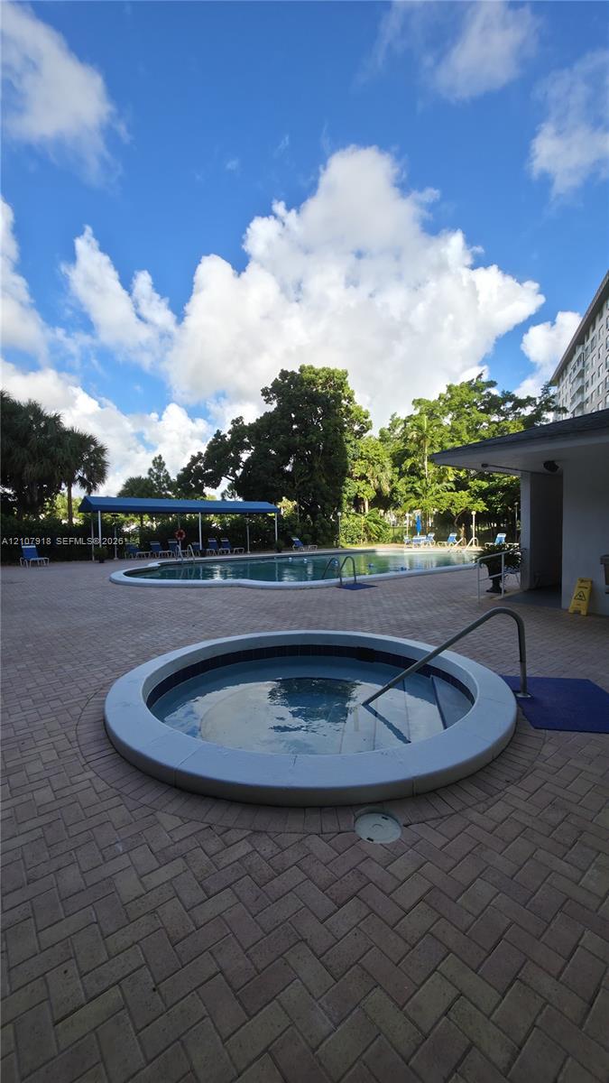 Resort-style outdoor recreation area featuring a circular spa with integrated seating, expansive paved terrace with herringbone brick pattern, mature landscaping with established trees, covered pavilion structure, and adjoining pool facility with blue canopy shade elements