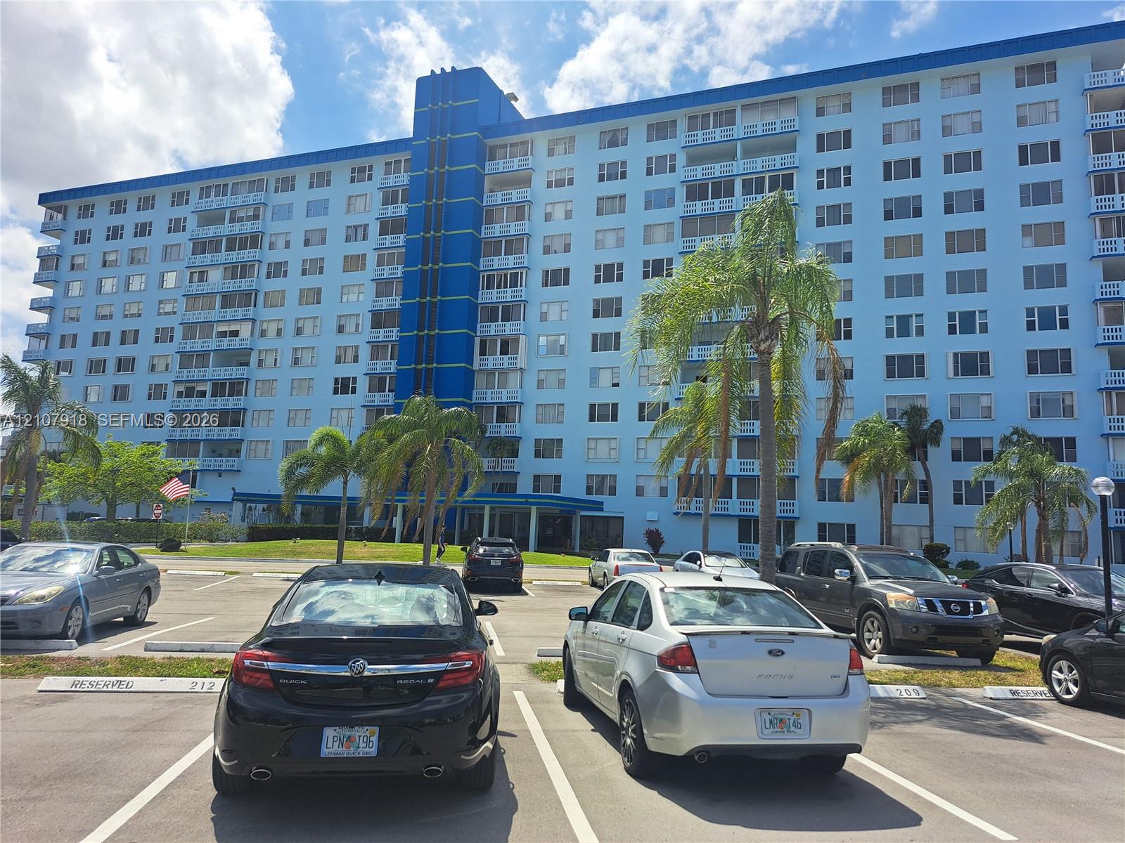 Multi-story residential building featuring a blue exterior facade, private balconies, and dedicated parking area with mature palm trees Multi-story residential building featuring a blue exterior facade, private balconies, and dedicated parking area with mature palm trees