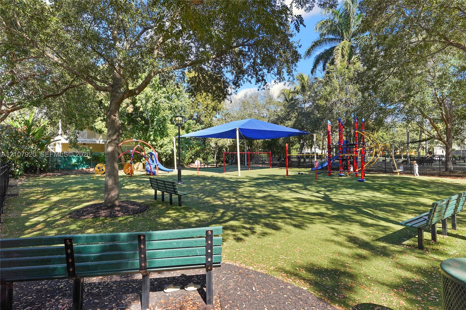 Recreational outdoor space featuring permanent playground structures, a large shade canopy, mature shade trees, and built-in metal bench seating