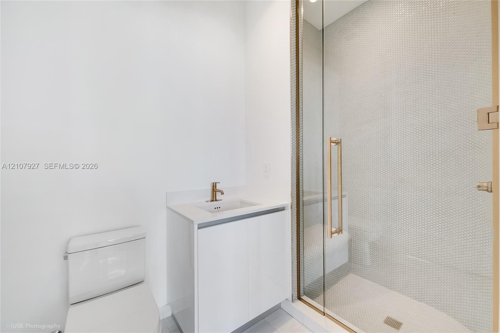 Modern bathroom featuring a glass-enclosed shower with small-format tile surround, a minimalist vanity with a white countertop and gold-tone faucet, and a sleek, low-profile toilet