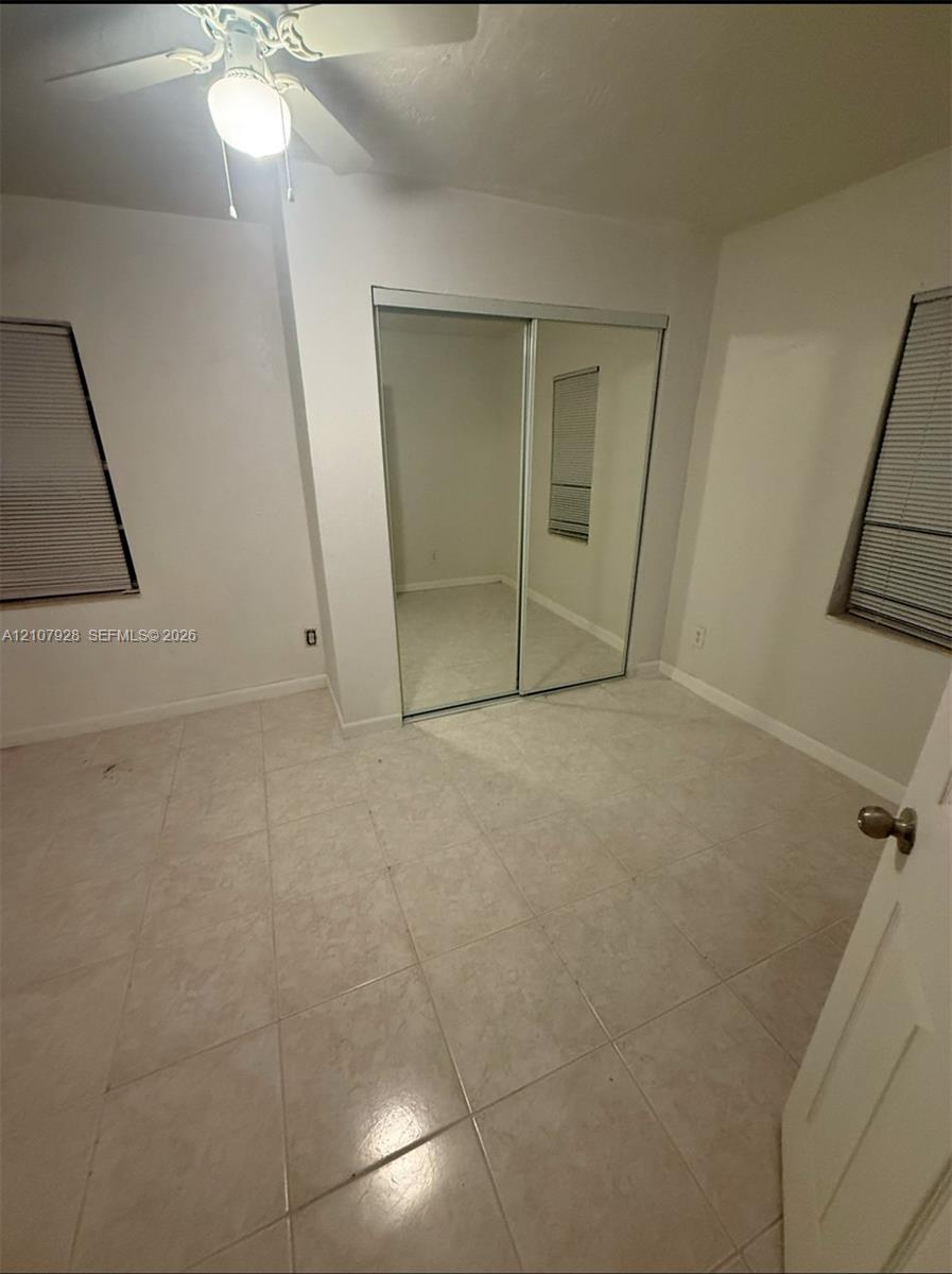 Neutral tile flooring, sliding mirrored closet doors, dual windows with fitted blinds, and a central ceiling fan with an integrated light fixture