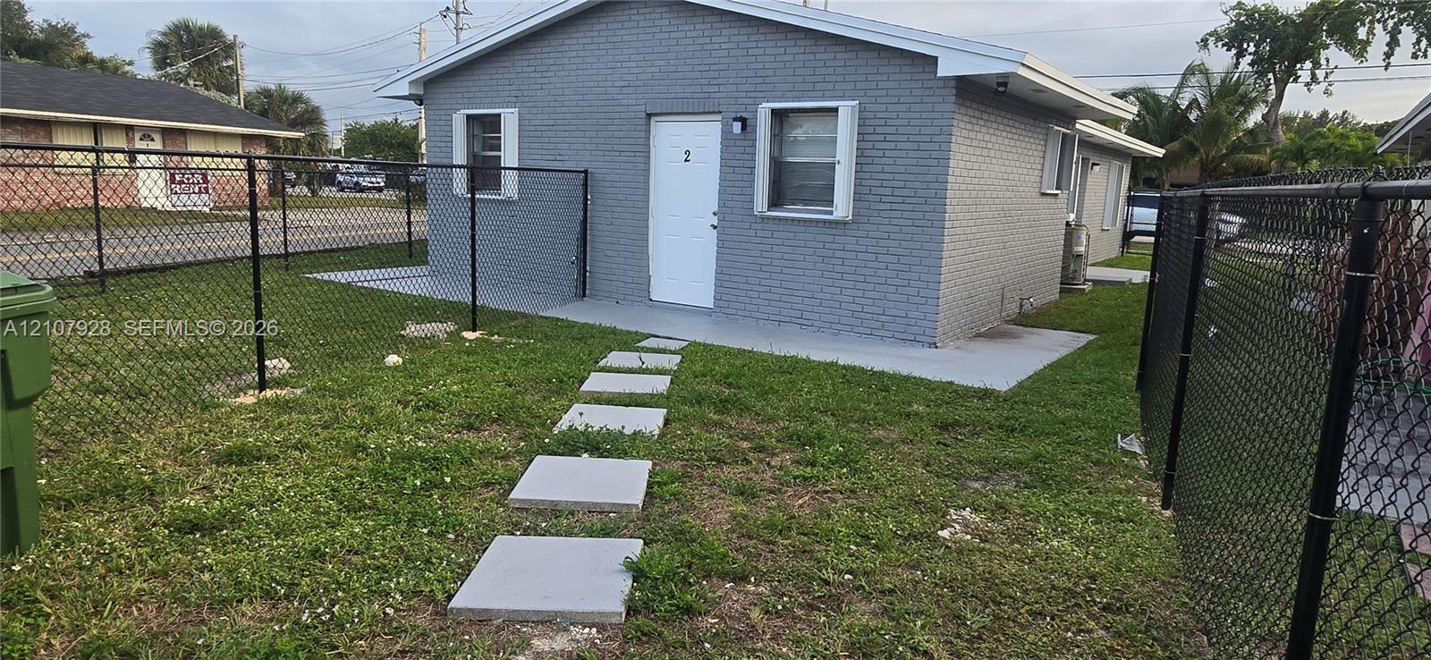 Painted brick exterior featuring a concrete entry pad, square paver walkway, and black chain-link perimeter fencing
