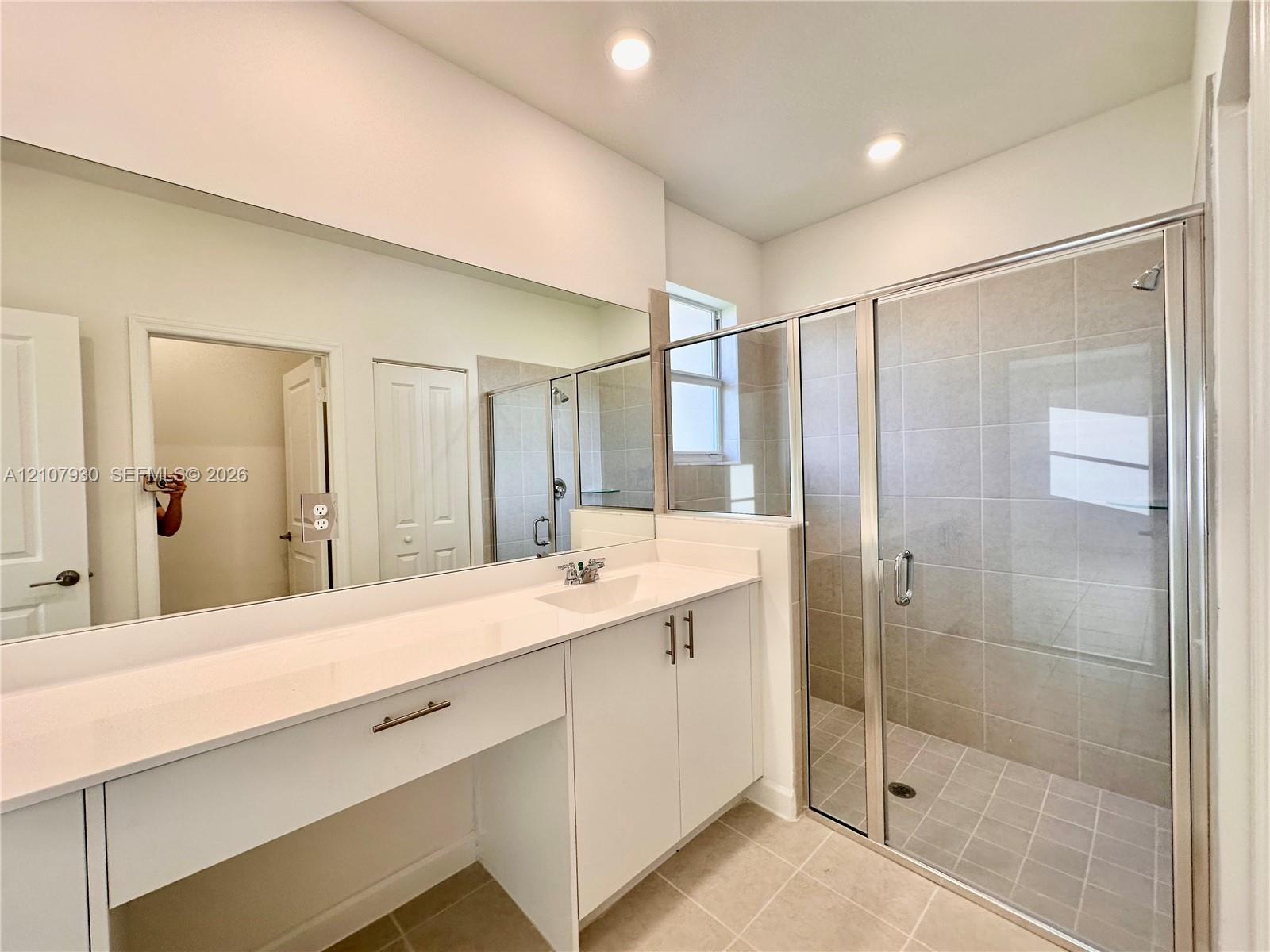 Bathroom featuring a white vanity with a solid-surface countertop, an integrated sink, full-width vanity mirror, walk-in shower with tile surround, and recessed ceiling lighting