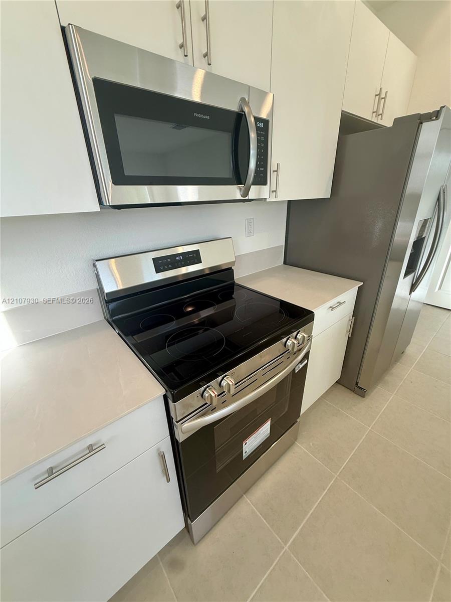 Kitchen featuring flat-panel white cabinetry, stainless steel microwave, glass-top electric range, stainless steel refrigerator, and neutral-toned floor tile
