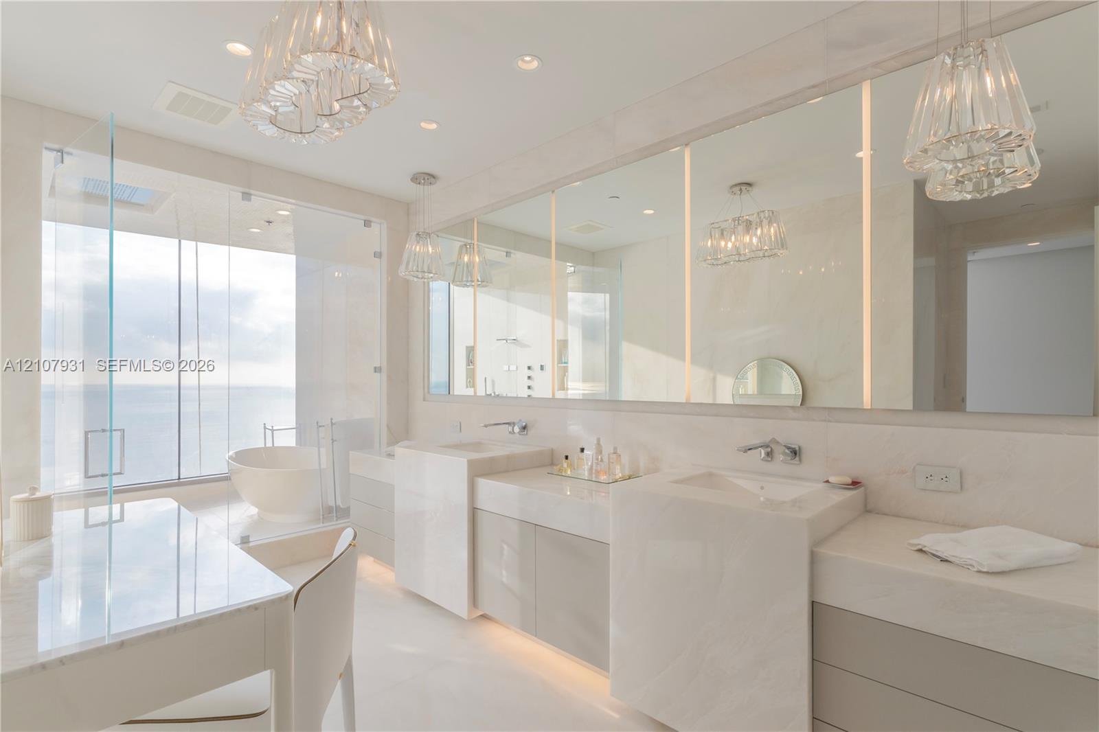 Dual-vanity cabinetry with stone-finish countertops, integrated soaking tub behind floor-to-ceiling glass, expansive wall-to-wall mirror, and tiered glass pendant lighting