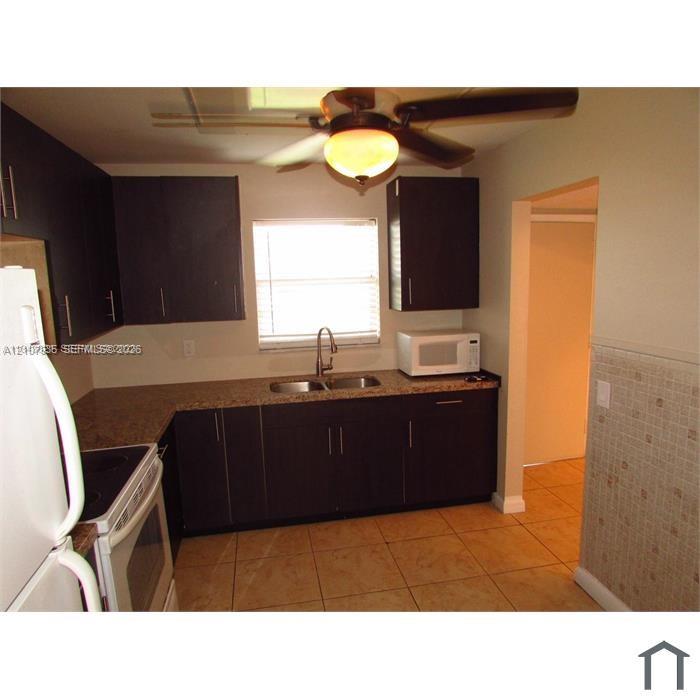 Kitchen featuring dark-toned cabinetry, stone-finish countertops, stainless steel dual-basin sink with arched faucet, ceramic tile flooring, and ceiling fan with integrated lighting