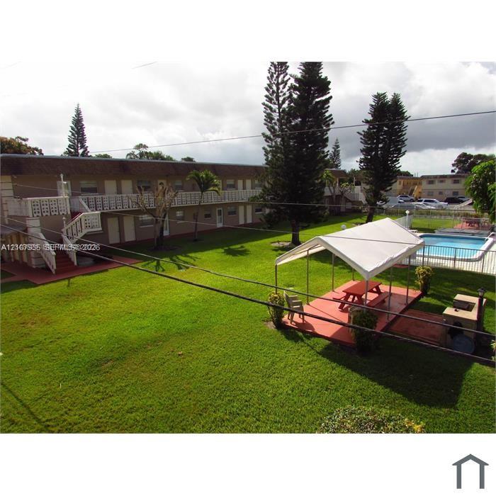 Expansive common area featuring a manicured lawn, mature trees, a swimming pool with perimeter fencing, and a paved patio with a built-in barbecue station