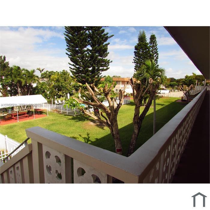 Elevated exterior balcony featuring decorative breeze block railing overlooking a shared greenspace, mature landscaping, and an enclosed outdoor swimming area