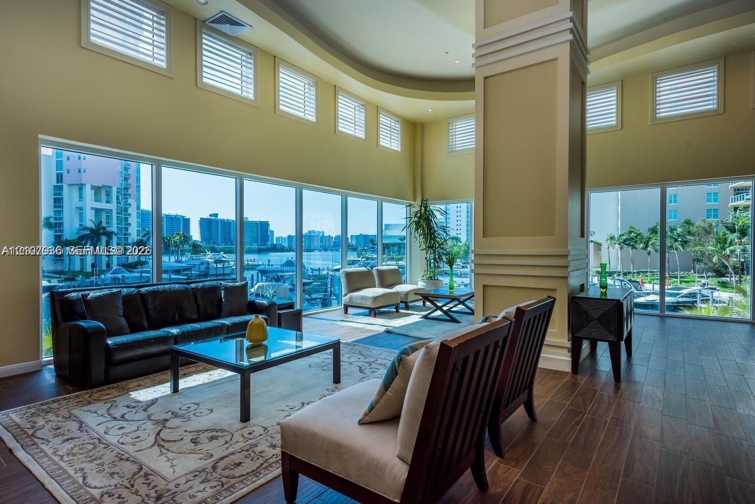 Expansive living space featuring floor-to-ceiling windows, soaring tray ceilings, decorative millwork on a prominent support column, and wood-finish flooring