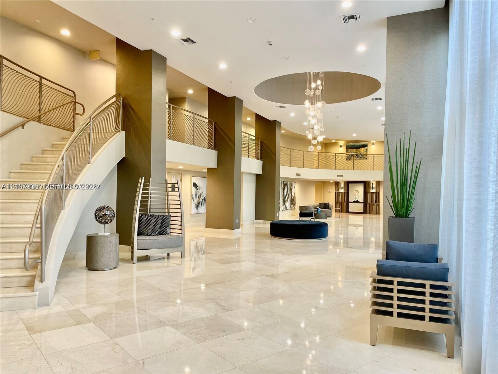 Grand lobby featuring polished stone flooring, a sweeping staircase with custom metal railing, multi-level mezzanine, and a recessed ceiling with a contemporary pendant chandelier