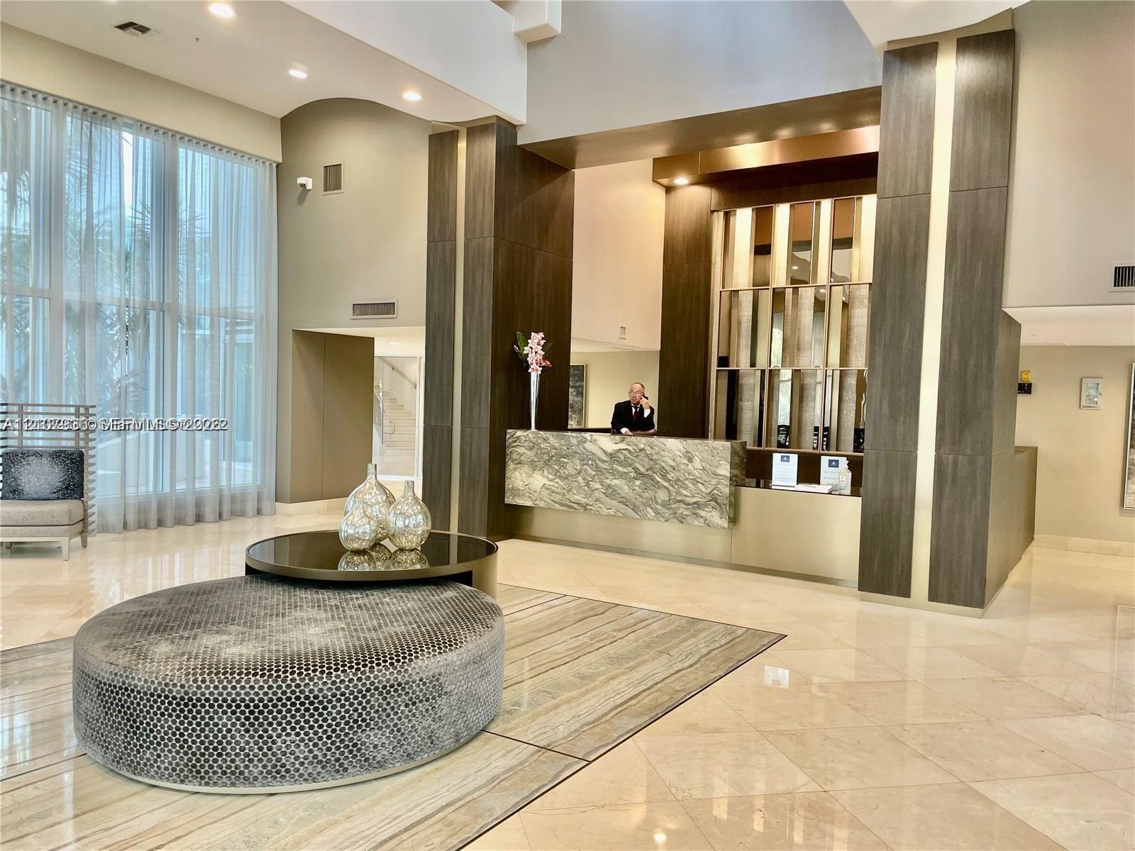 Double-height lobby featuring floor-to-ceiling windows, polished stone flooring, and a decorative stone-front reception desk accented with metallic paneling and dark wood-finish columns