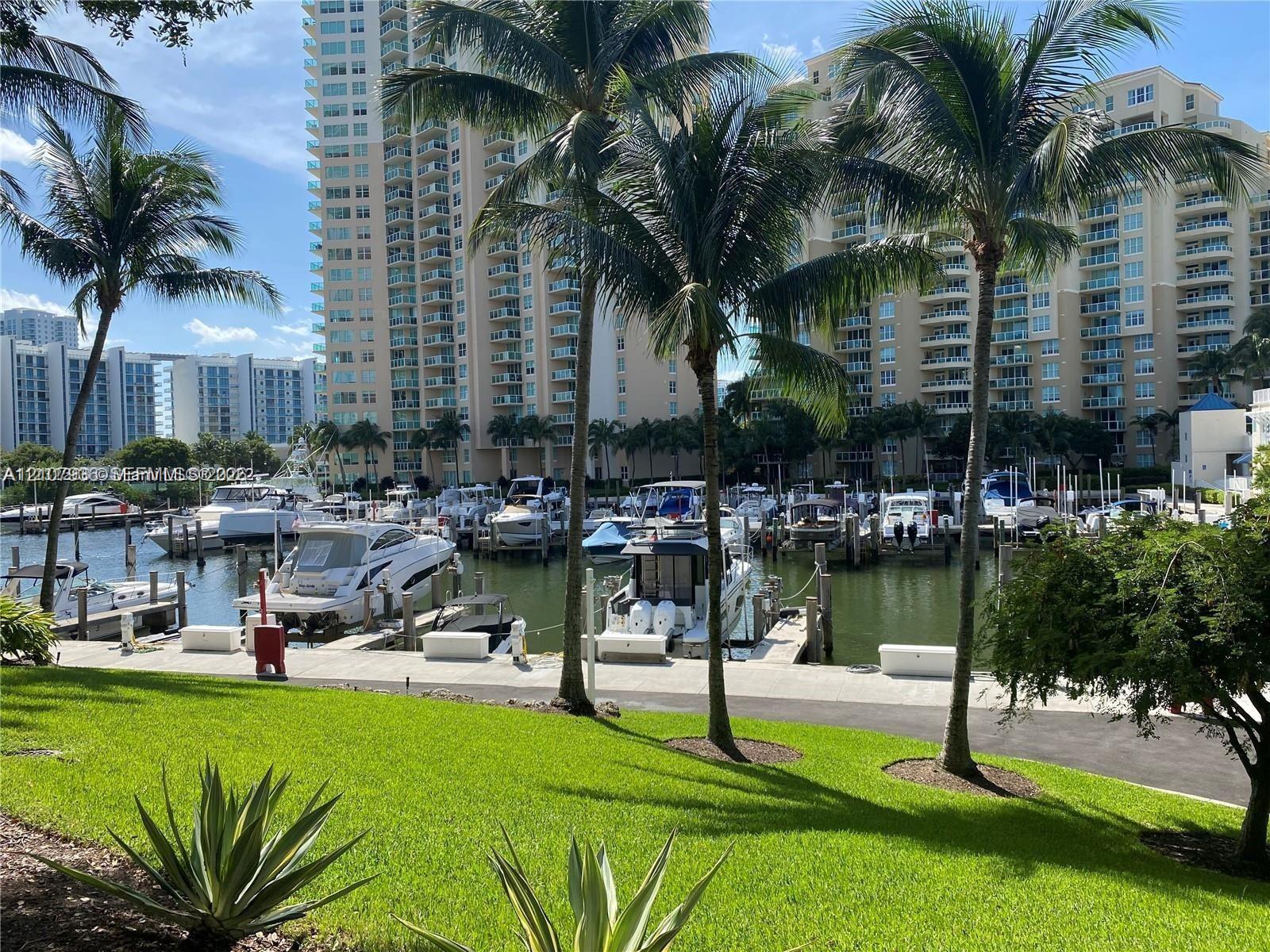 Marina featuring multiple boat slips, paved waterfront walkway, and manicured green space with mature palm trees