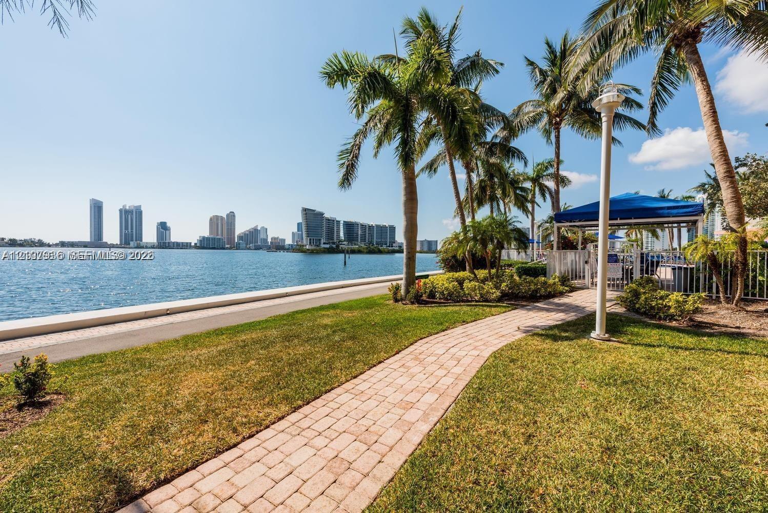 Waterfront grounds featuring a brick paver pathway, mature palm trees, coastal landscaping, and perimeter fencing