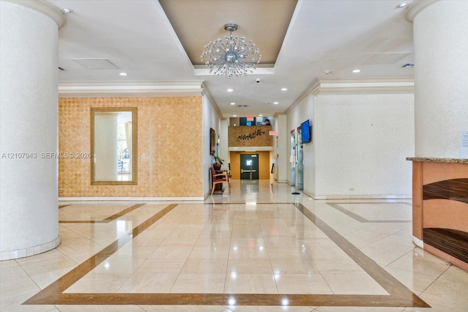 Grand lobby featuring polished stone tile flooring with decorative inlay, a central coffered ceiling with a crystal chandelier, structural circular columns, and a textured stone accent wall with an integrated mirror