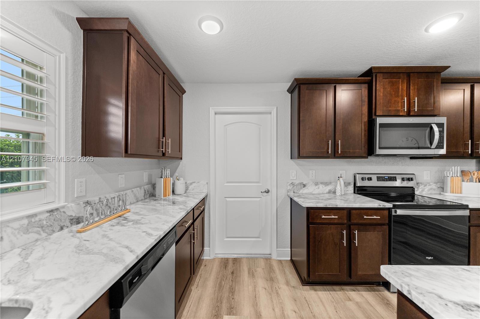 Kitchen featuring stained wood cabinetry, stone-patterned countertops, stainless steel appliances, wood-finish flooring, and shutter-style window treatments