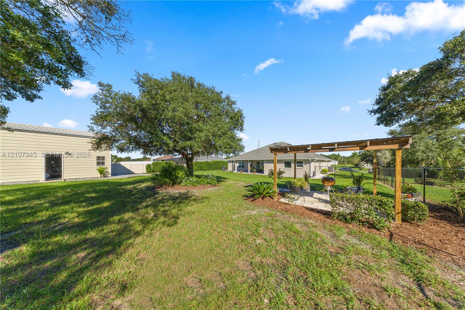 Expansive backyard featuring a wood pergola, stone-paver patio, mature shade trees, and a detached structure with light-colored siding