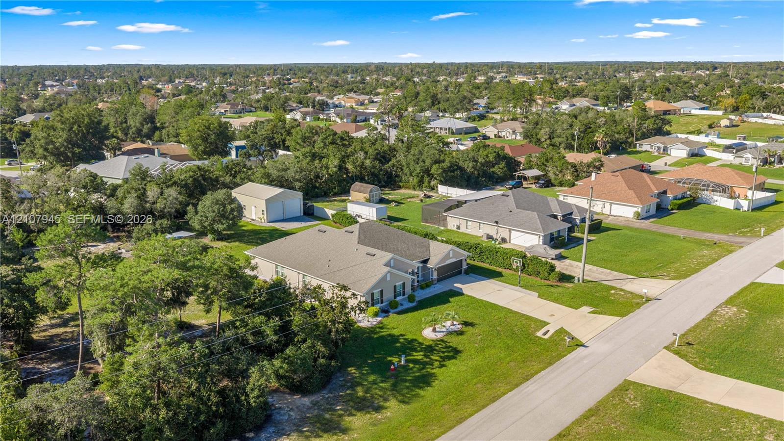 Aerial view of a property featuring a single-story residence, a multi-car paved driveway, a detached metal-sided storage structure, and expansive grounds with mature trees