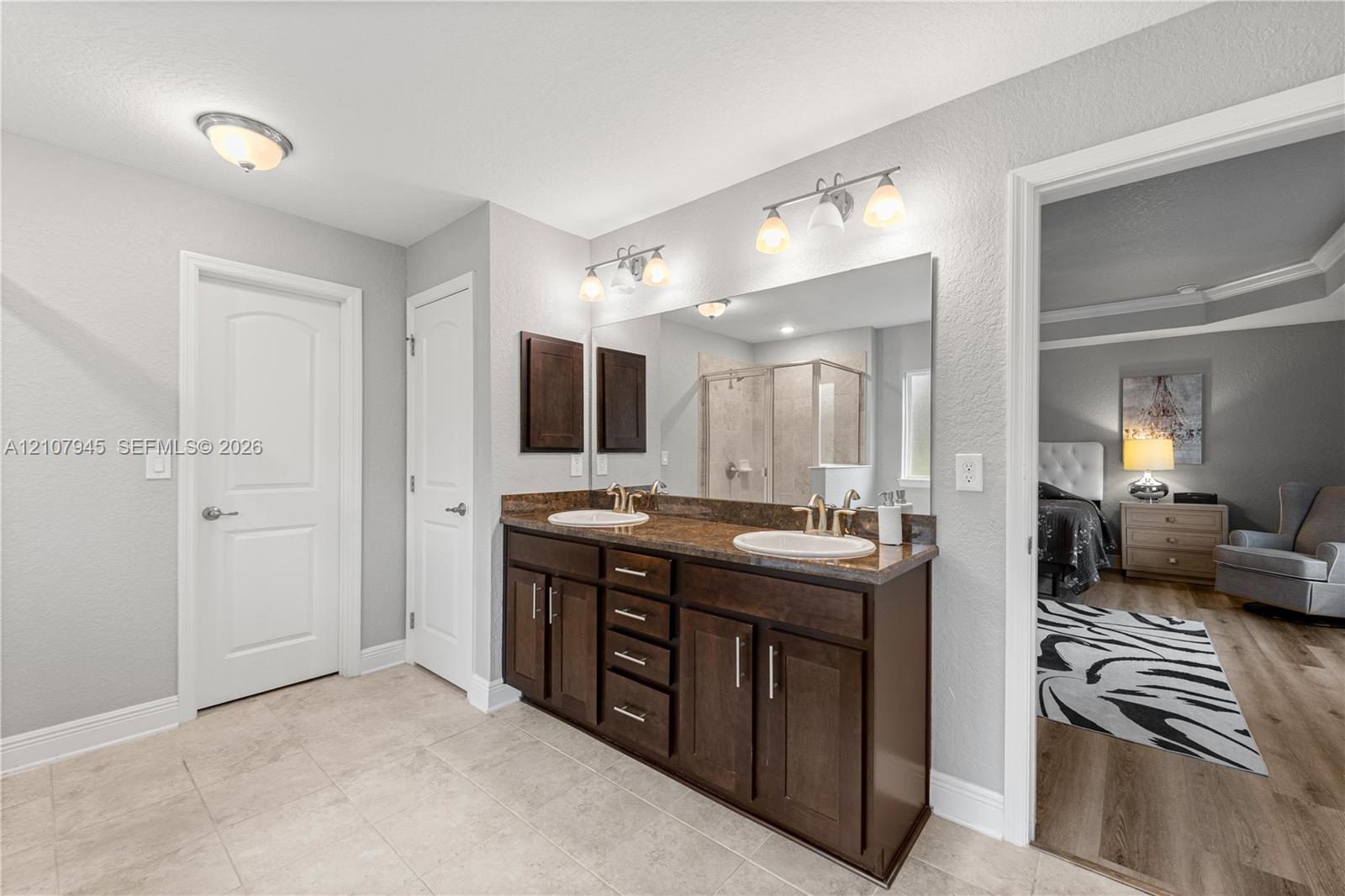 Dual-sink vanity with dark wood cabinetry, stone-finish countertop, full-width vanity mirror, and tile flooring