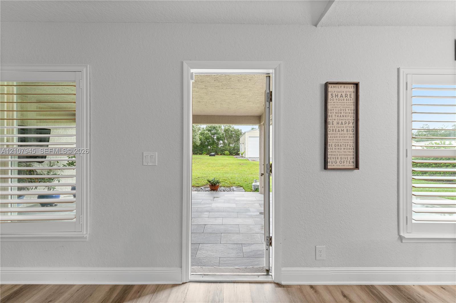 Interior featuring wood-finish flooring, dual windows with plantation-style shutters, and an exterior door opening to a paved patio area