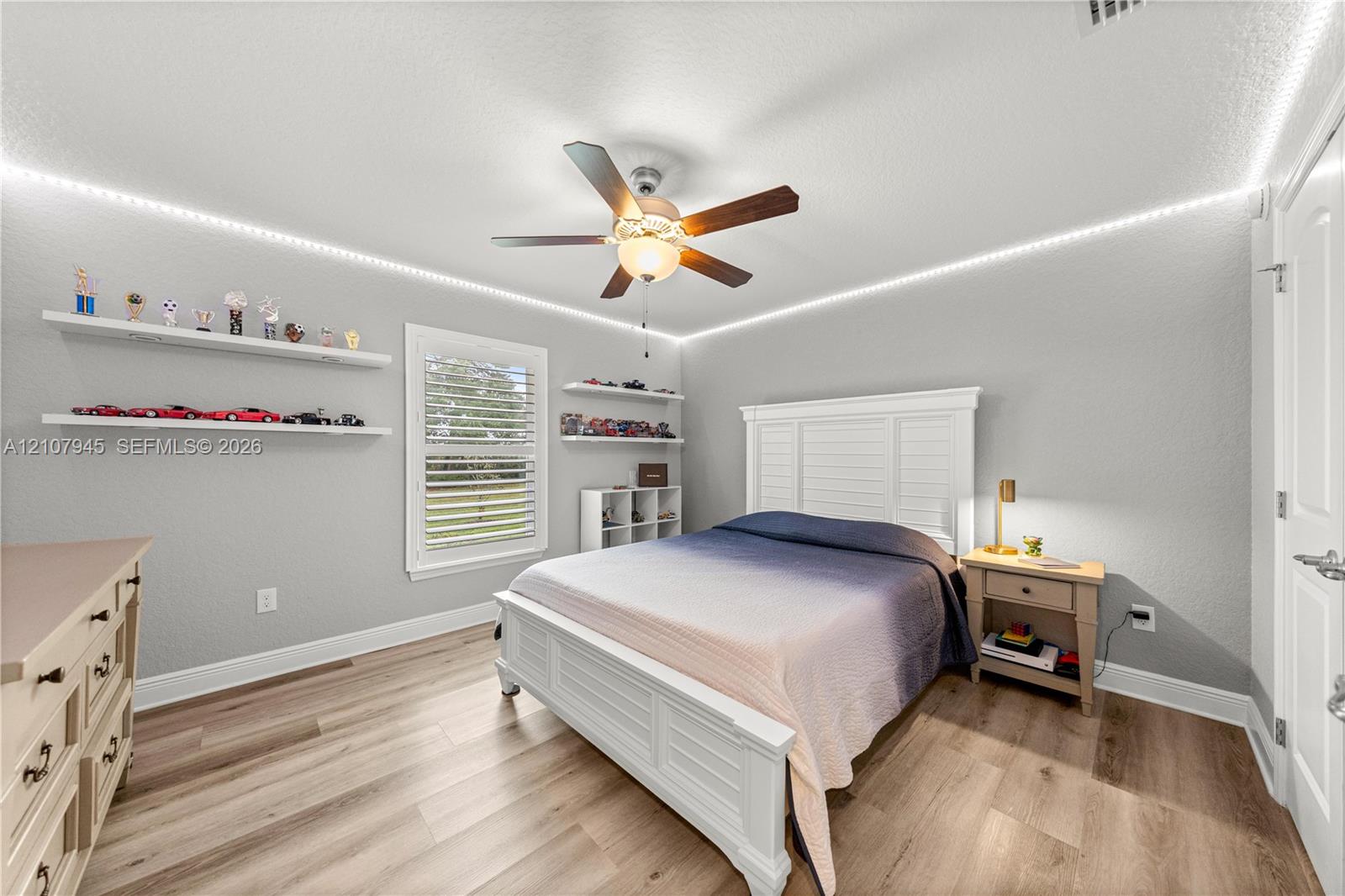 Wood-finish flooring, ceiling fan with integrated lighting, window with shutter treatments, and installed wall-mounted shelving