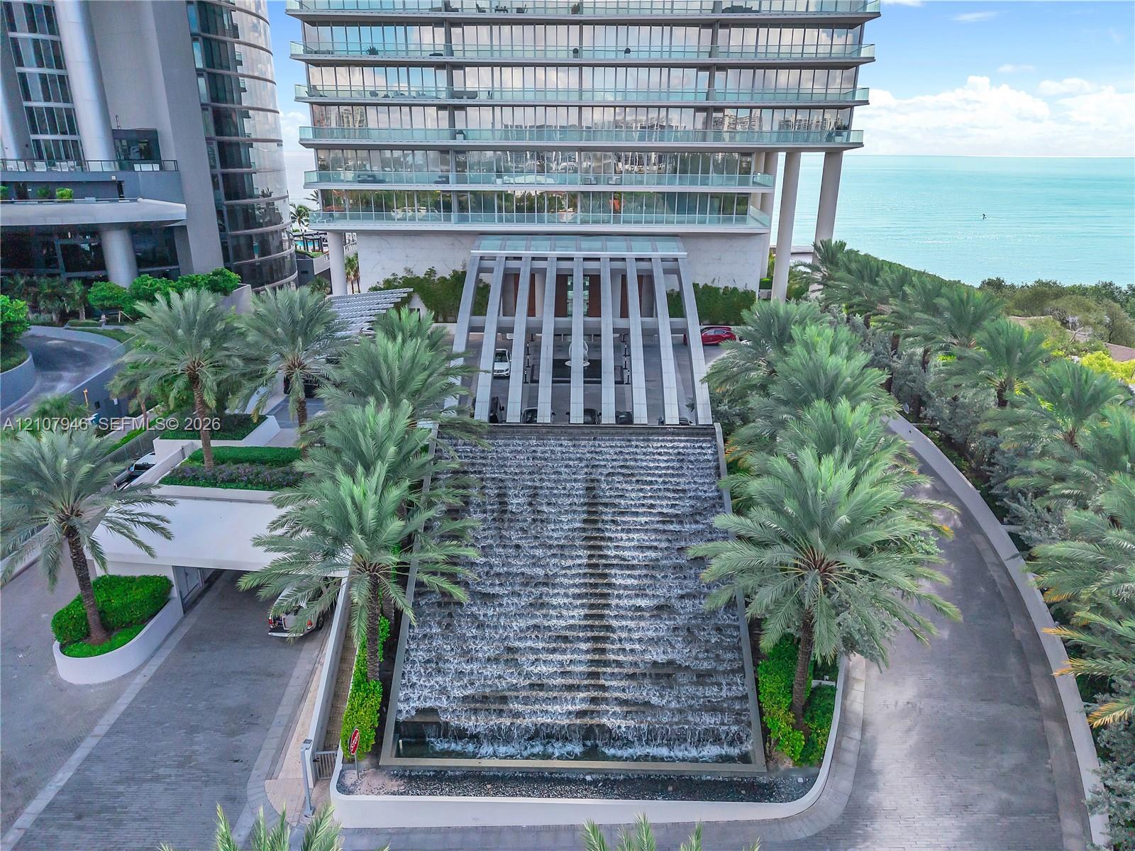 High-rise residential building featuring a tiered waterfall feature, a covered porte-cochre with structural pillars, landscaped palm trees, and glass-railed balconies