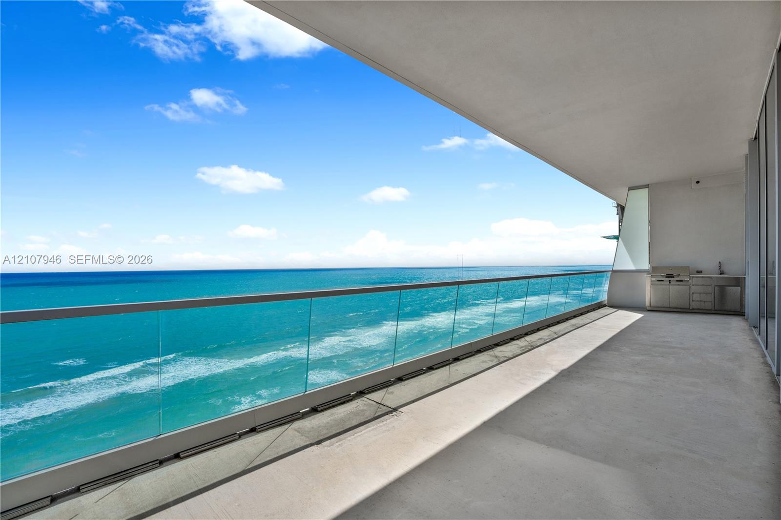 Expansive balcony featuring a glass panel railing, concrete flooring, and an outdoor kitchen area with stainless steel cabinetry and a built-in grill
