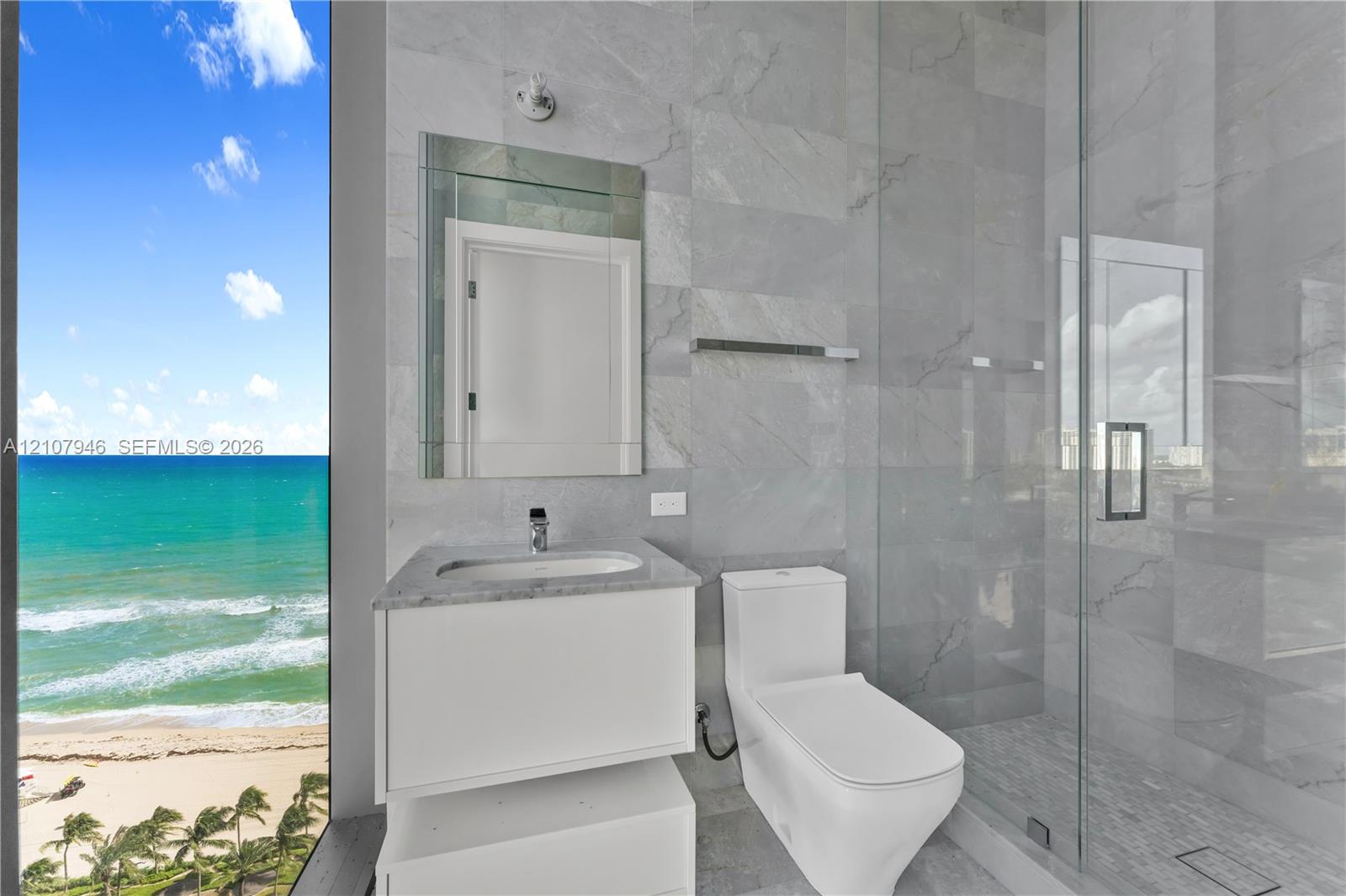 Floor-to-ceiling glass panel, wall-mounted vanity with a stone countertop, frameless glass-enclosed shower, large-format tile wall cladding, and oceanfront scenery