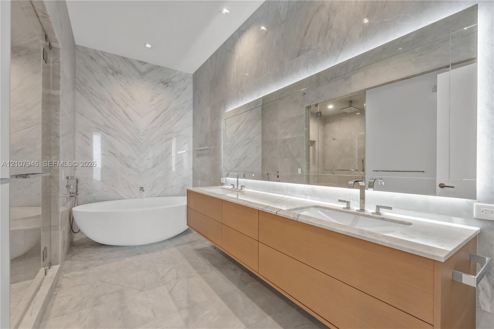 Floating wood-finish vanity with dual integrated sinks, stone countertop, back-lit wall mirror, and freestanding soaking tub set against full-height stone wall tile