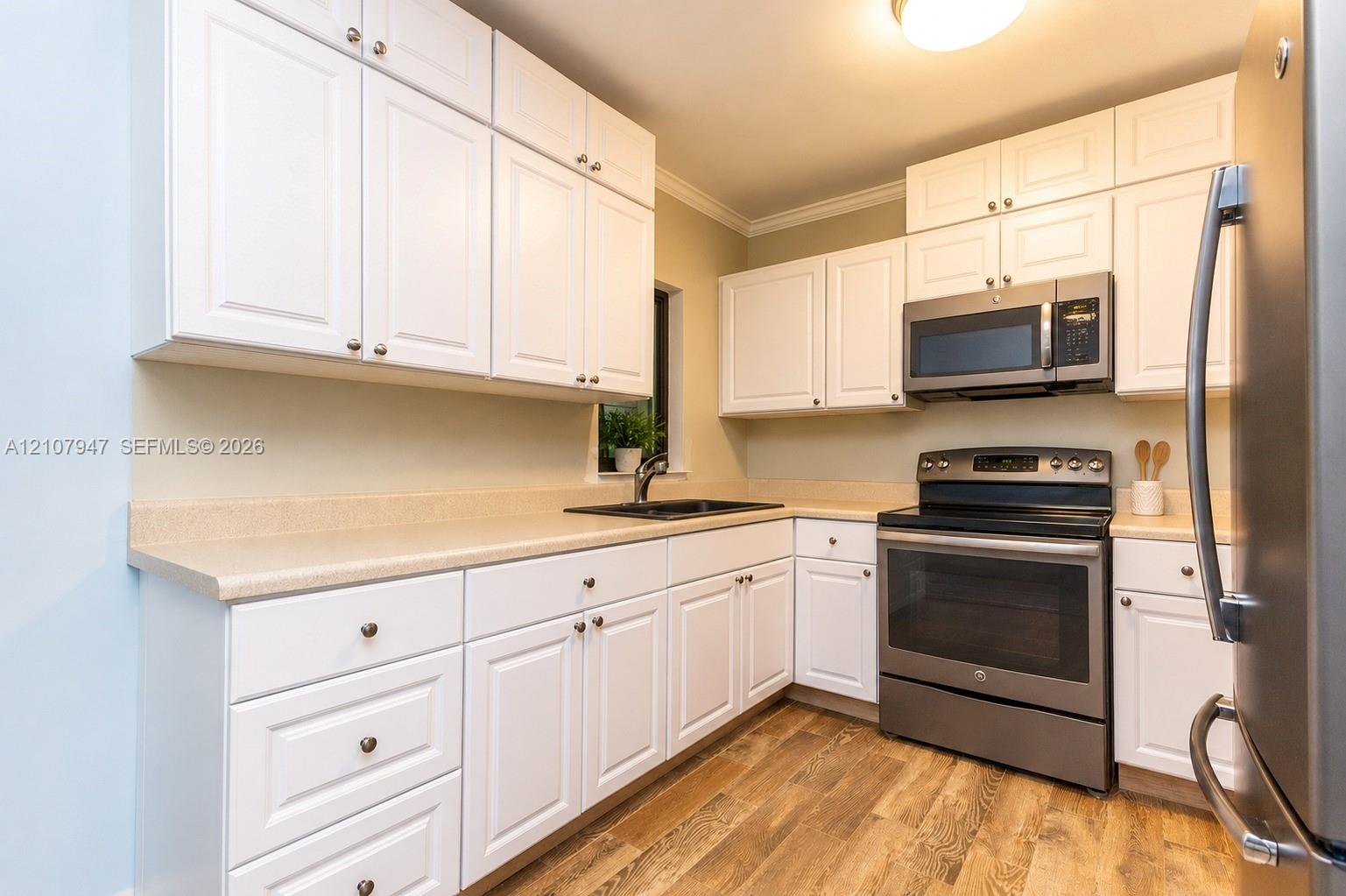 Kitchen featuring white shaker-style cabinetry, solid-surface countertops, wood-finish flooring, and stainless steel appliances Kitchen featuring white shaker-style cabinetry, solid-surface countertops, wood-finish flooring, and stainless steel appliances