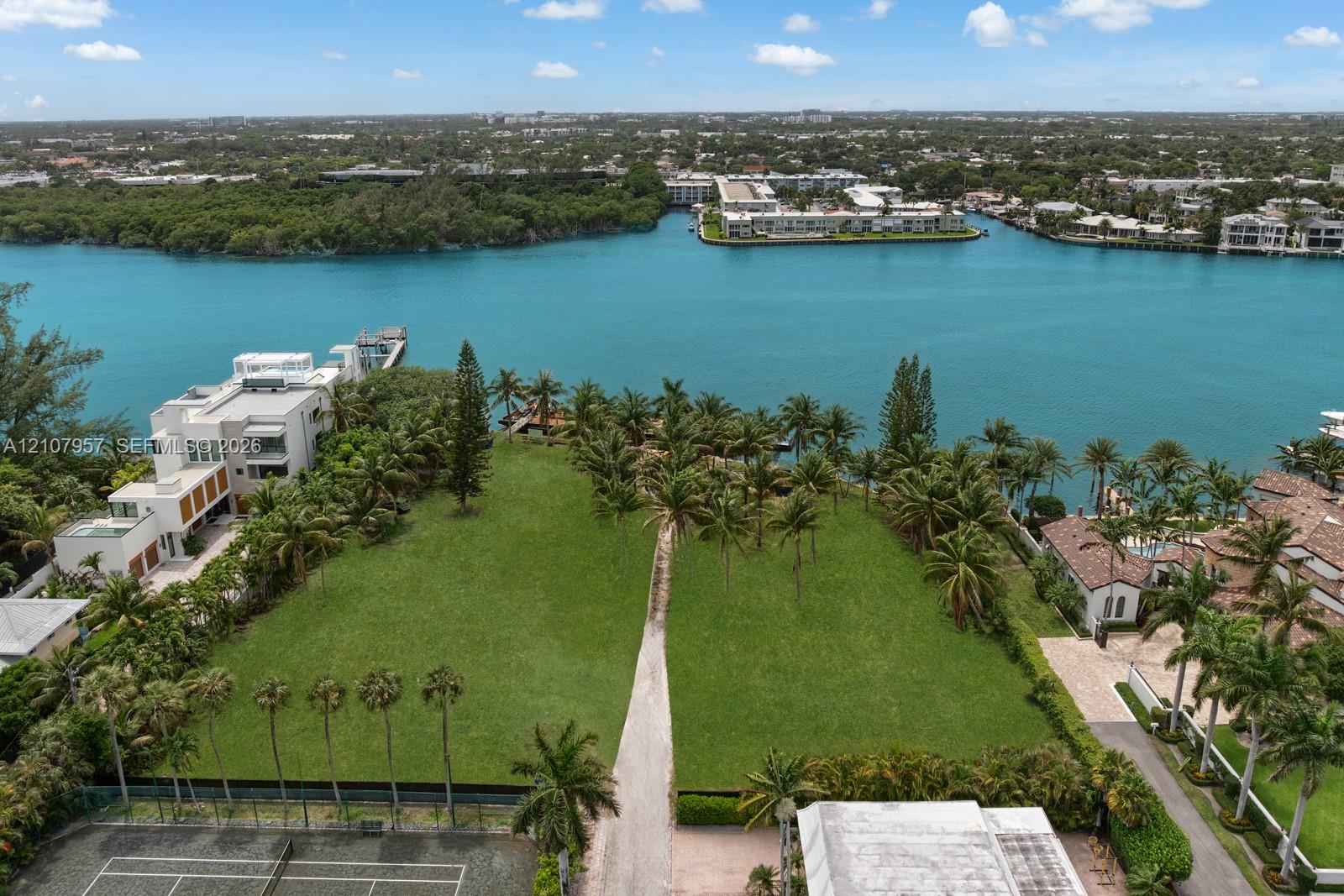 Waterfront lot featuring direct canal access, expansive manicured lawn, mature palm trees, and a hard-surface tennis court