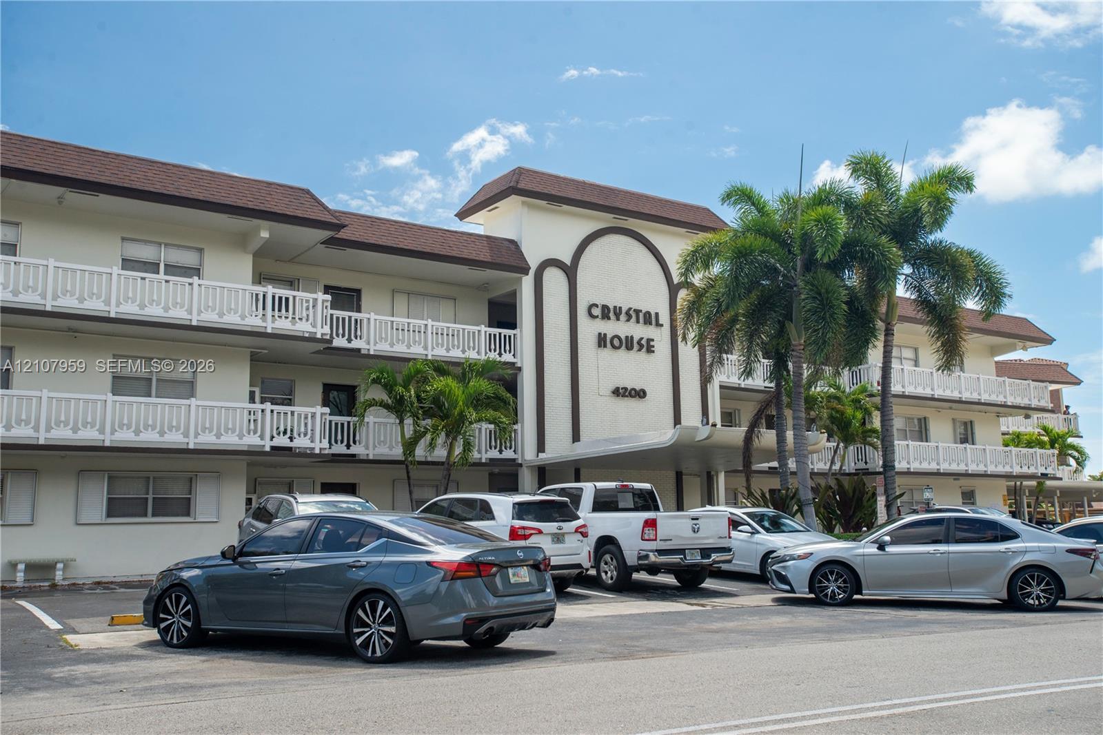 Multi-story exterior featuring a shingled roof, white balcony railings, a covered entrance portico, and integrated mature palm landscaping