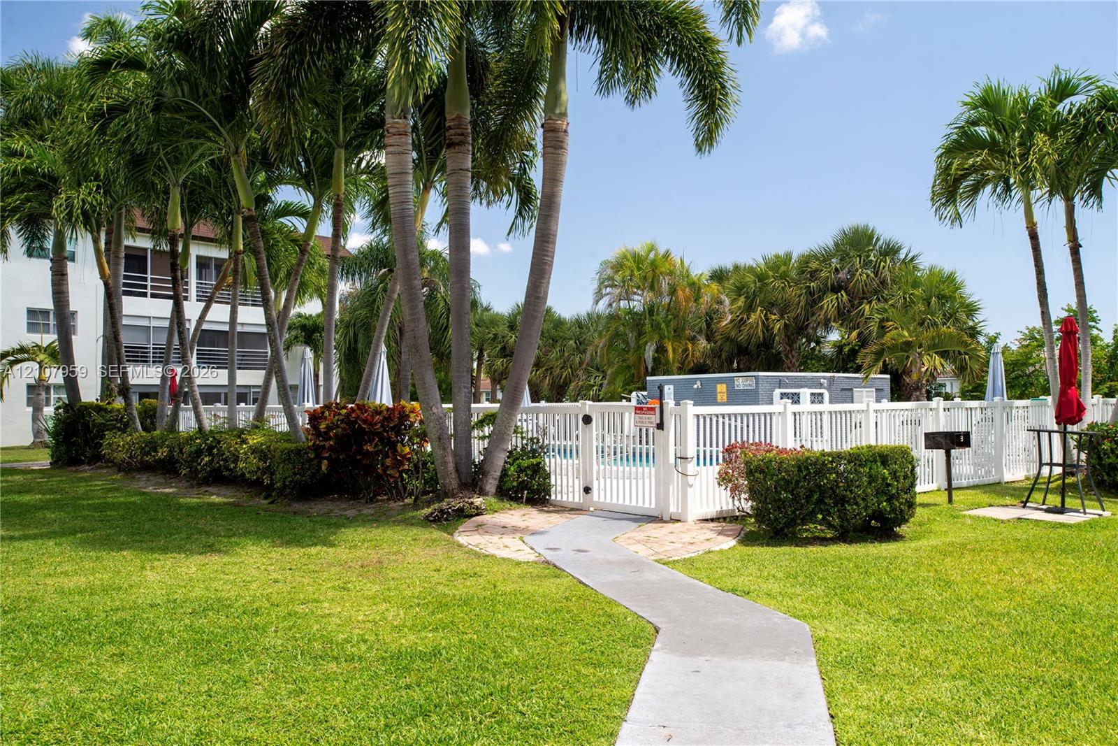 Swimming pool area featuring a white vinyl perimeter fence, concrete paver pathway, manicured lawn, and mature palm trees