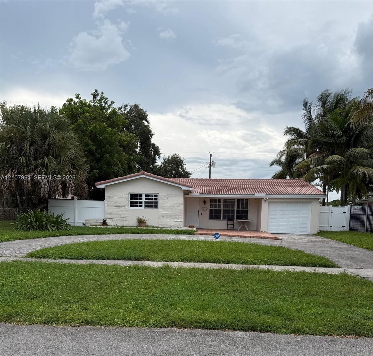 Single-story residence featuring a terracotta-tiled roof, textured stucco facade, attached garage, paved circular driveway, and white vinyl perimeter fencing