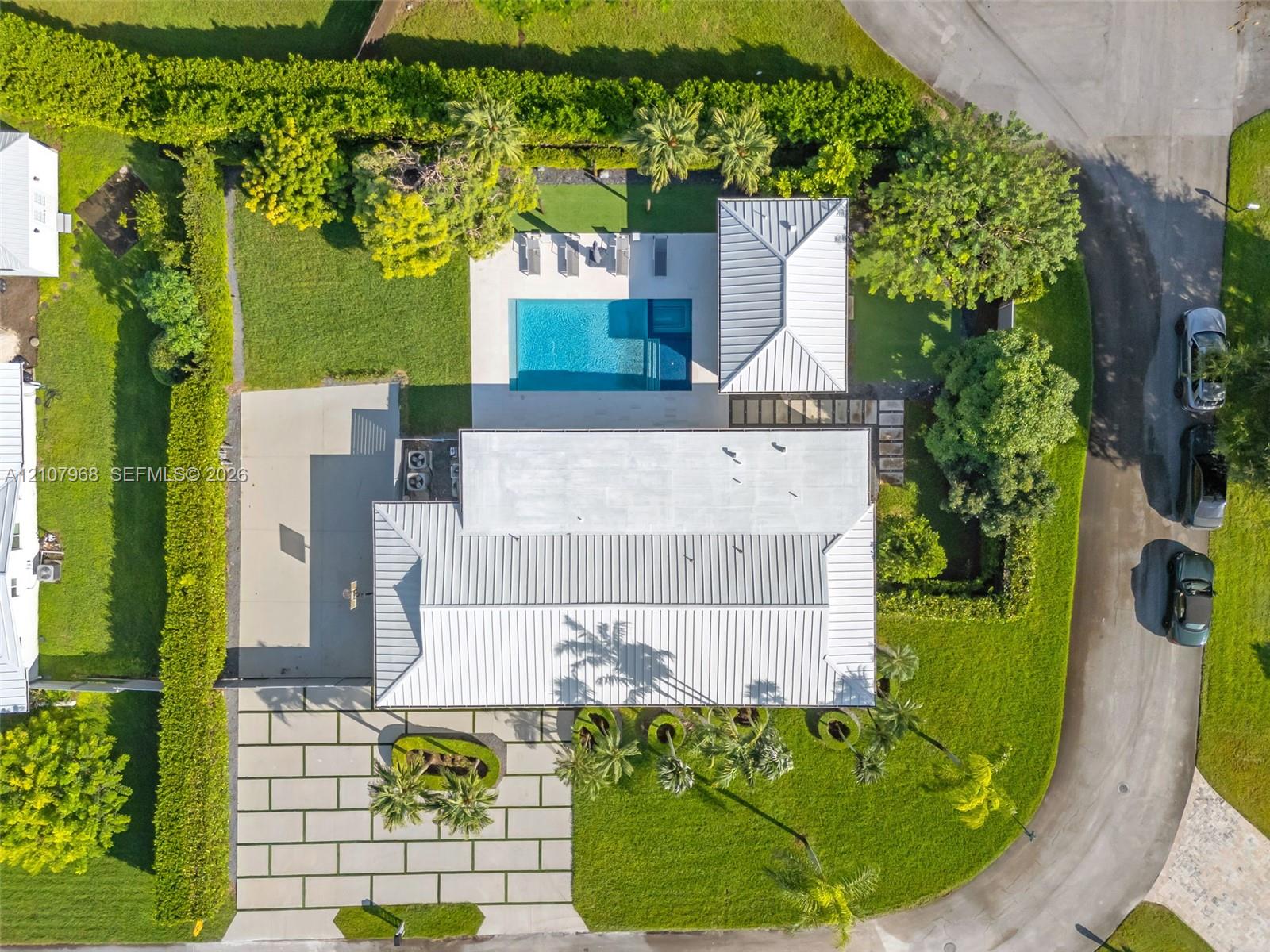 Aerial view of property featuring a metal roof, rectangular swimming pool, expansive concrete paver driveway, manicured lawn, and mature perimeter hedging