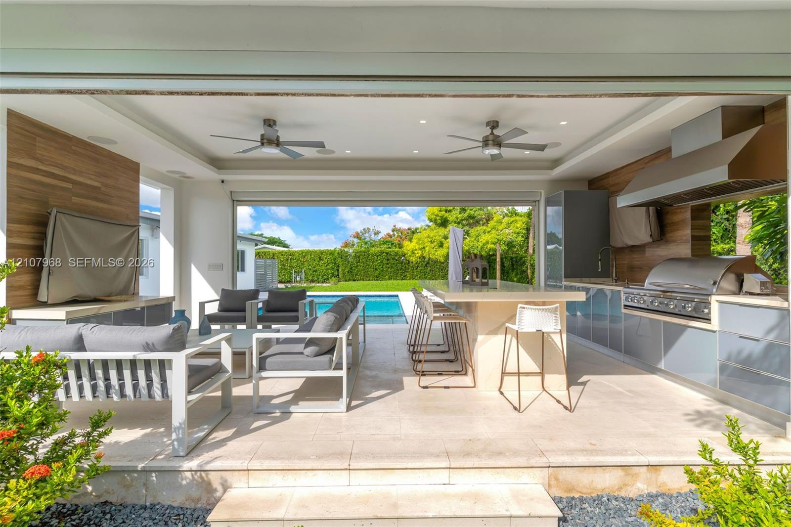 Covered outdoor living space featuring a built-in kitchen with a professional-grade stainless steel grill, stone-topped island with seating, wood-finish wall paneling, and integrated ceiling fans