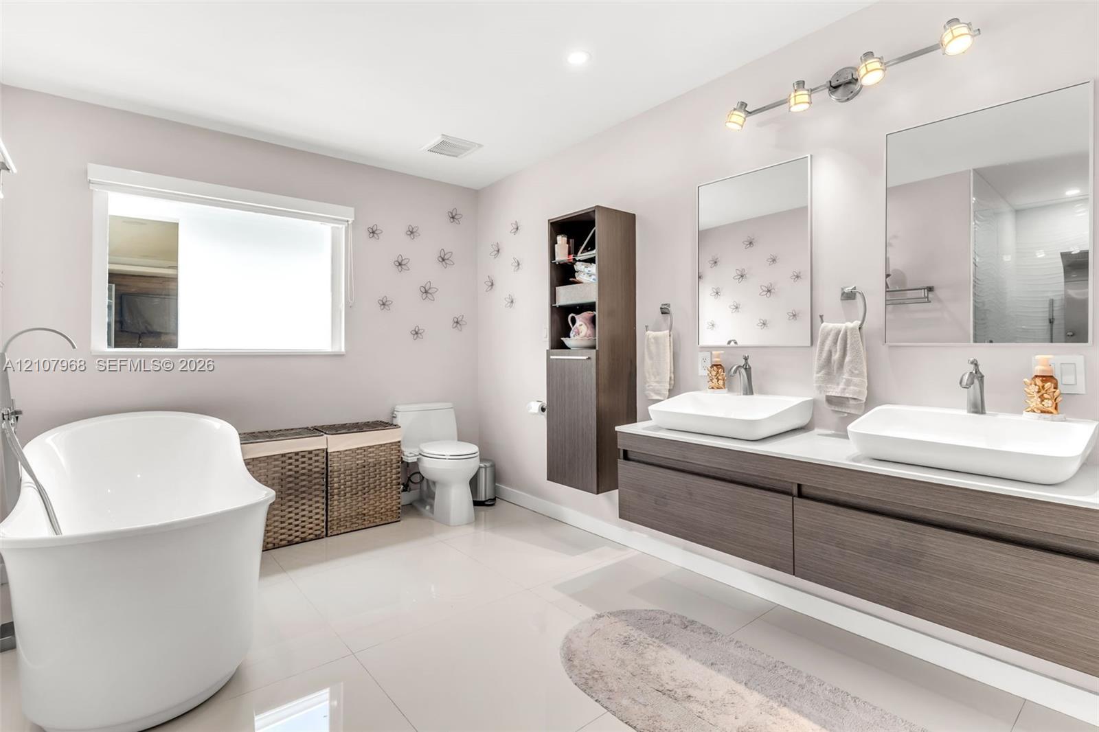 Floating vanity featuring wood-finish cabinetry, dual vessel sinks, and stone-surface countertop, complemented by a freestanding soaking tub, vertical built-in storage cabinet, and rectangular wall-mounted mirrors