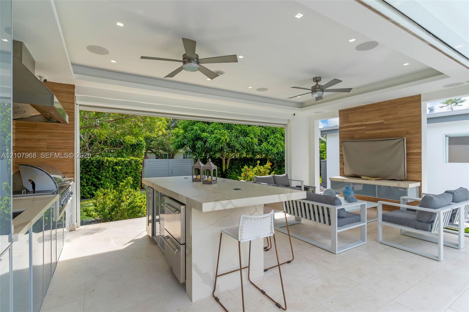 Covered outdoor kitchen and living space featuring a stone-topped island with stainless steel appliances, wood-finish wall paneling, dual ceiling fans, and large-format tile flooring