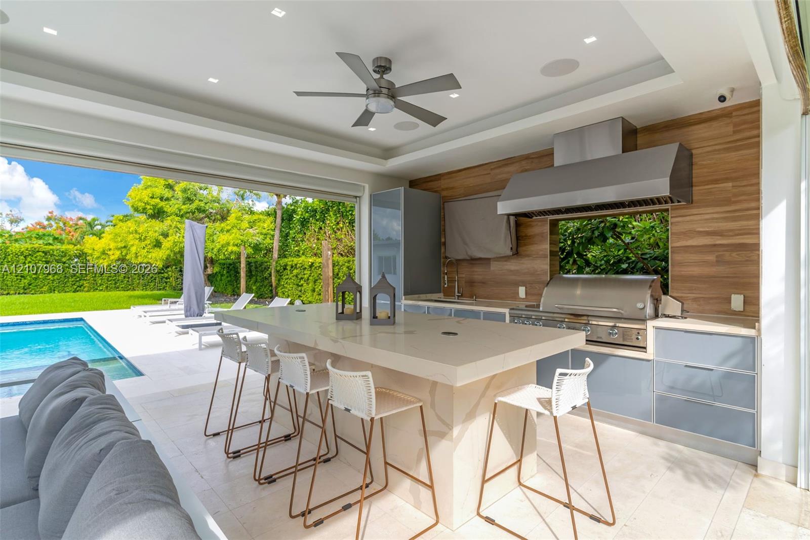 Covered outdoor kitchen featuring a stone-topped island, stainless steel grill, professional-grade range hood, wood-finish wall paneling, and recessed lighting