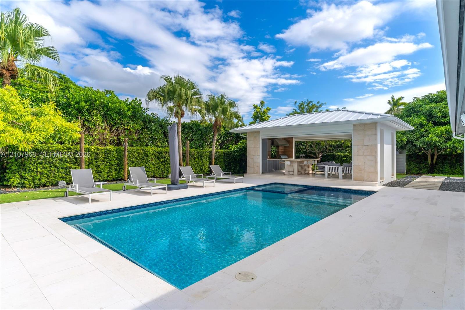 In-ground swimming pool with a stone patio, accompanied by a covered pavilion featuring a built-in outdoor kitchen and stone support columns