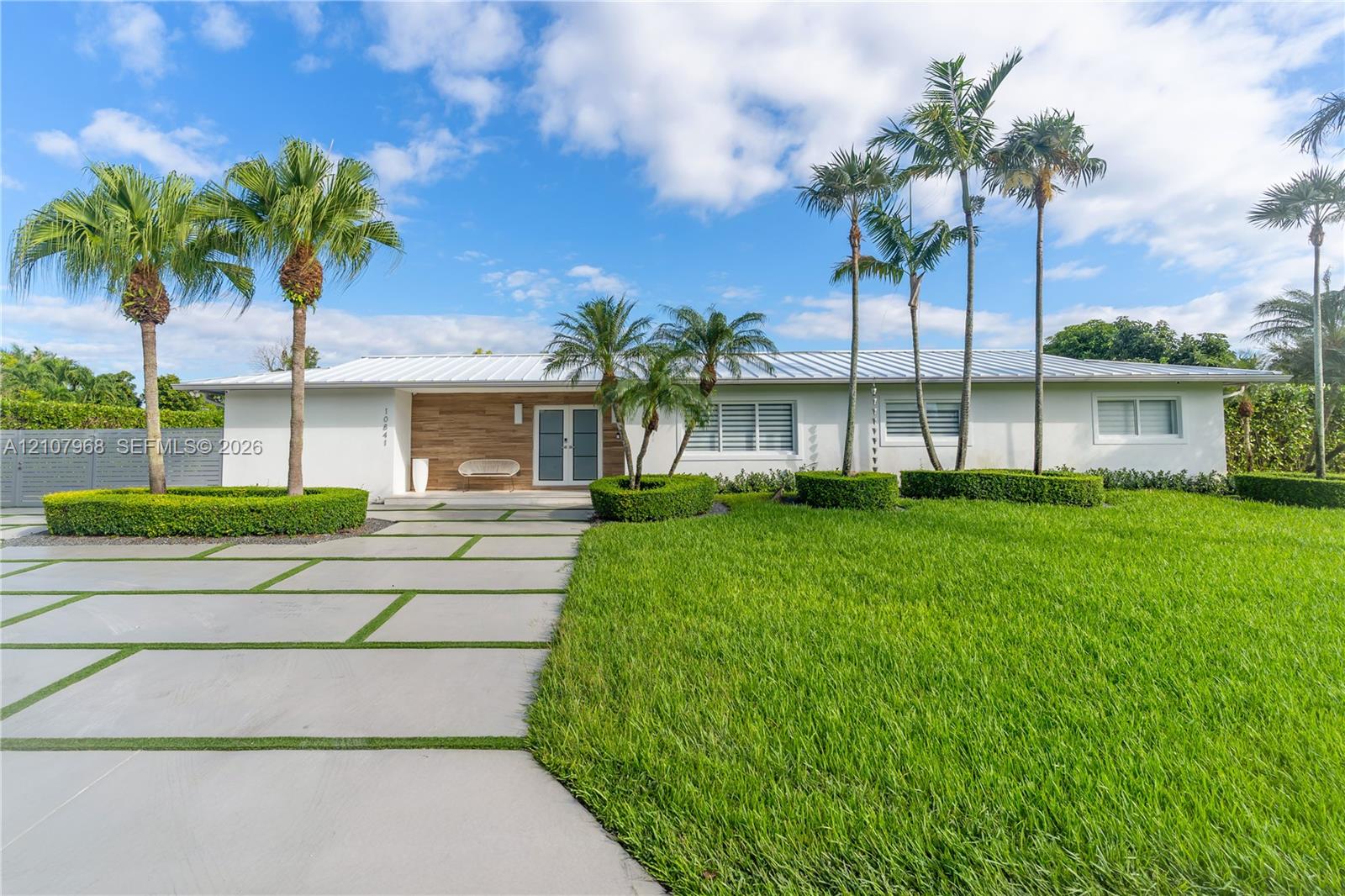 Single-story residence featuring a standing-seam metal roof, horizontal wood-panel siding, a concrete paver driveway with integrated turf, and professional landscaping with mature palm trees