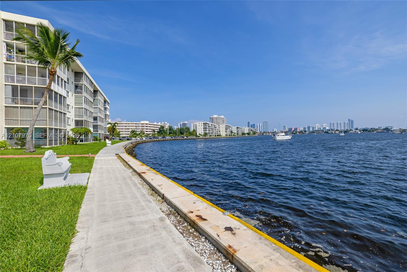 Waterfront multi-story residential building featuring screened-in balconies, concrete bulkhead, paved walkway, and manicured lawn