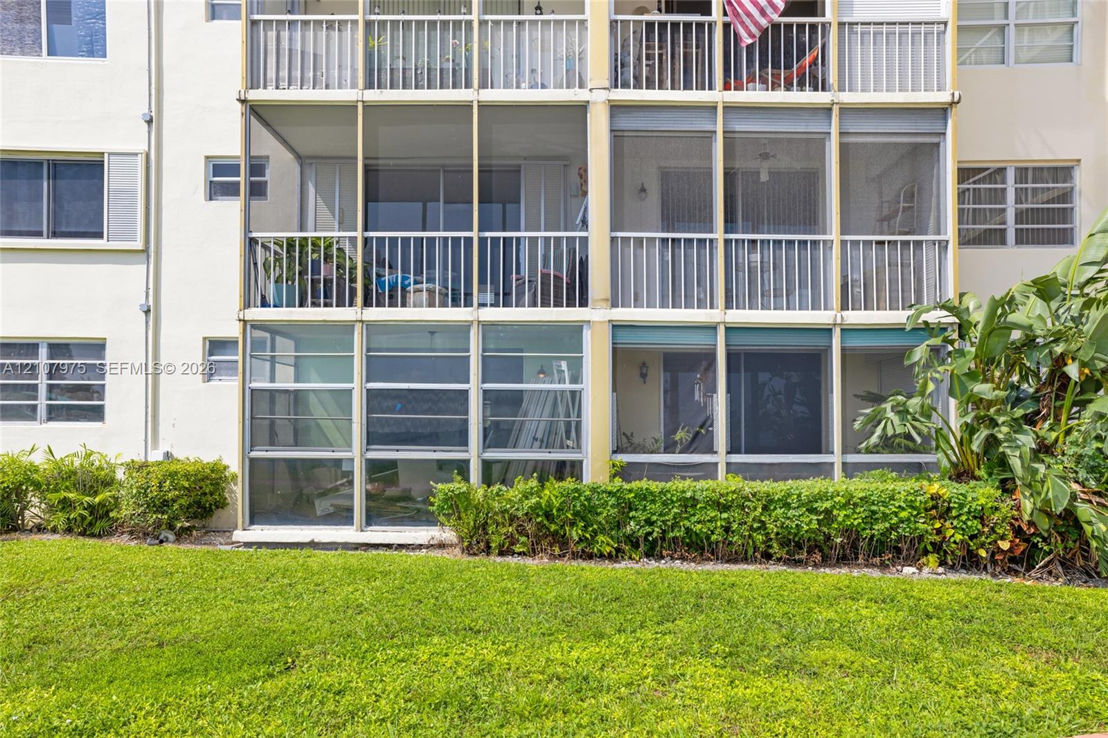 Multi-story building exterior featuring screened-in lanais, aluminum-frame balcony railings, and exterior windows with horizontal sliding panes, situated on a manicured lawn with mature landscaping