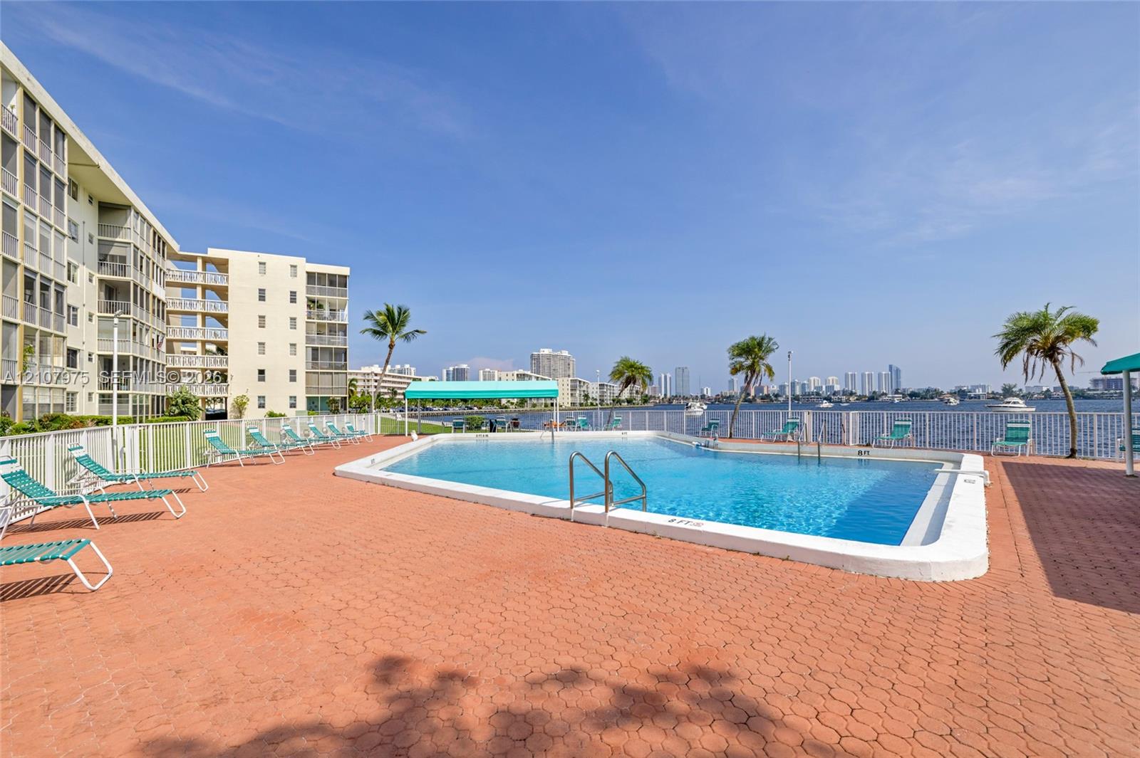 Outdoor swimming pool with integrated metal ladder, surrounding brick-paver deck, protective perimeter fencing, and established palm trees