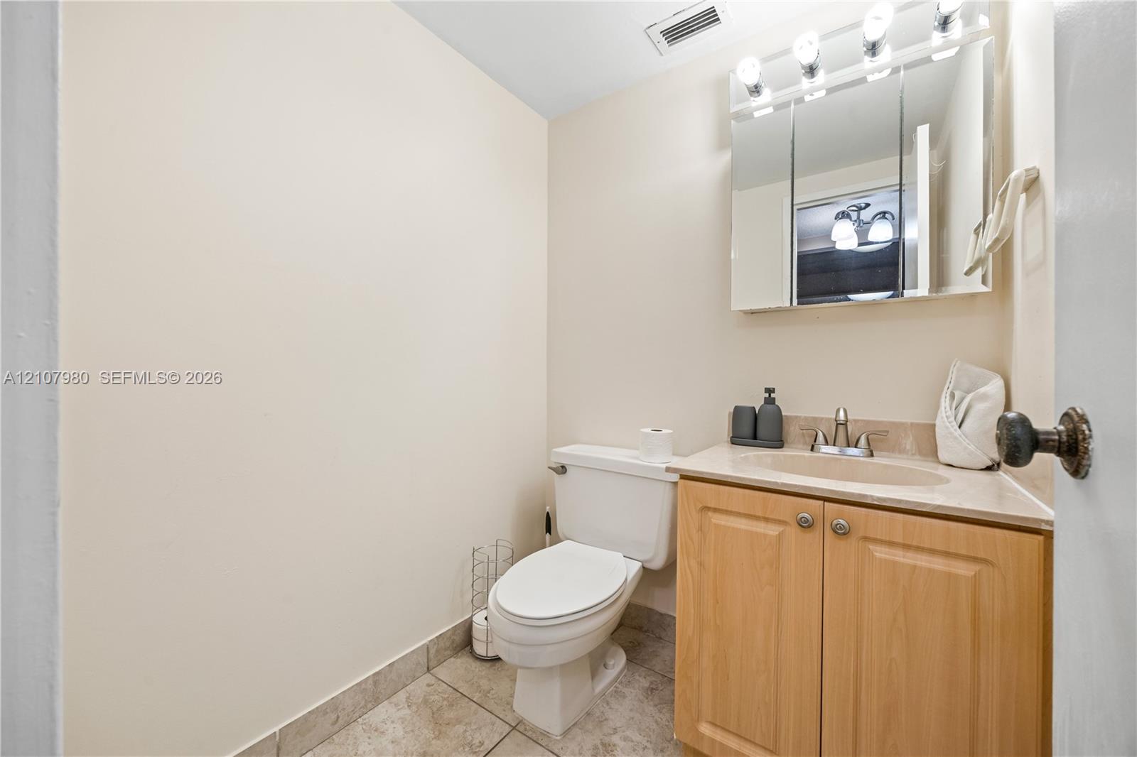 Vanity featuring light wood-finish cabinetry, a neutral-toned countertop with an integrated oval sink, a tri-panel mirrored medicine cabinet, and tile flooring