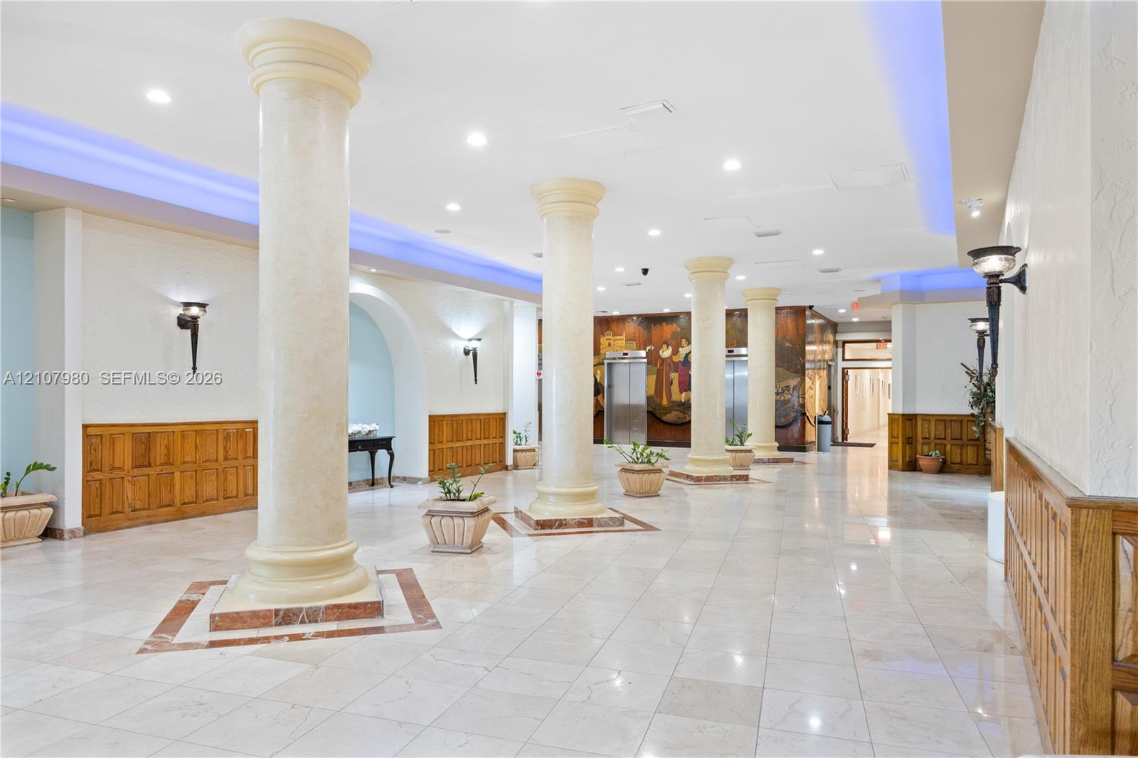 Grand lobby featuring polished stone flooring, architectural columns, wall-mounted sconces, wood-panel wainscoting, and metallic-finish elevator doors