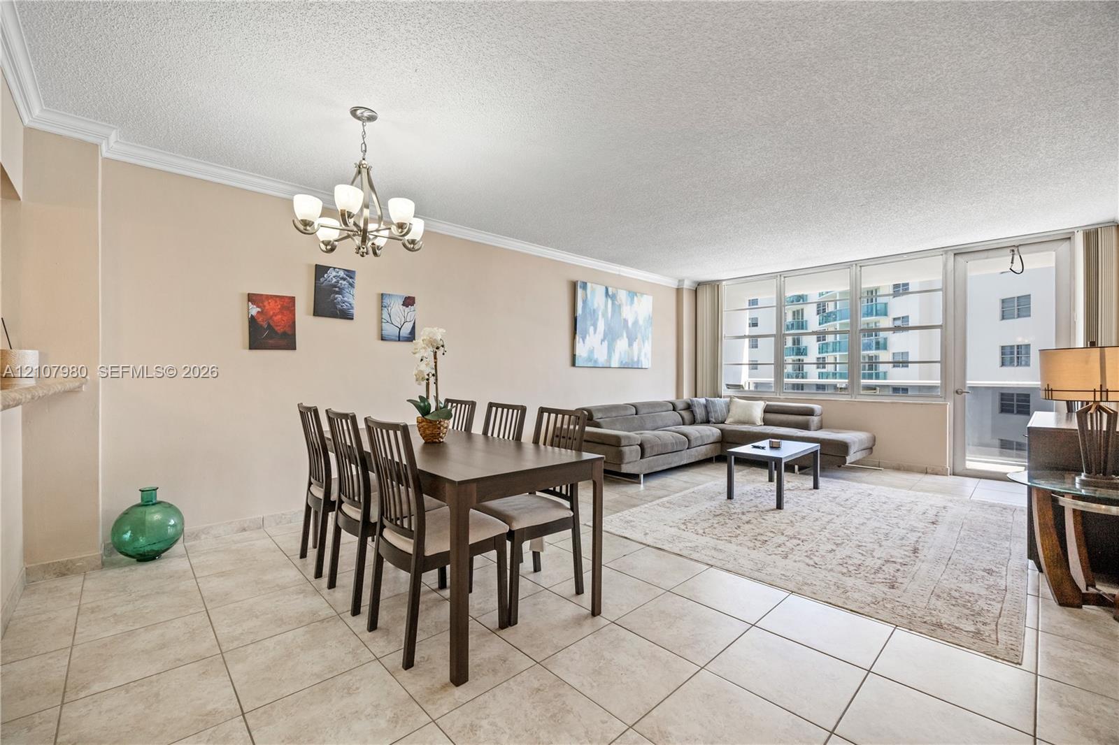 Open-concept living and dining area featuring large-format tile flooring, crown molding, multi-light chandelier, and floor-to-ceiling windows with a glass-paneled door