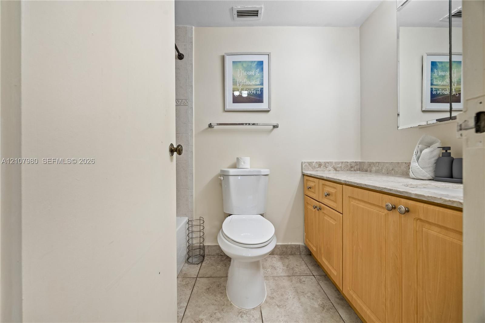 Bathroom featuring light wood-finish cabinetry, stone-style countertop, tile flooring, and a wall-mounted mirror