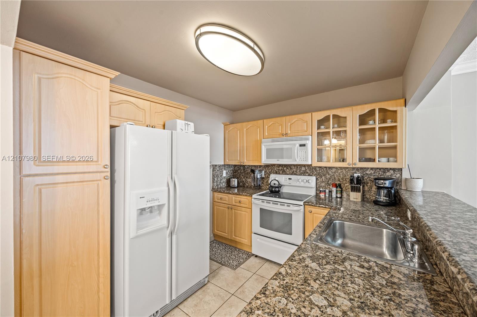 Kitchen featuring natural-finish wood cabinetry, stone-patterned countertops, a stainless steel undermount sink, glass-paneled upper cabinet doors, and white appliances
